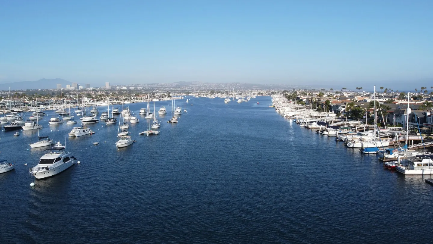 Sailboats and motorboats moored along Newport Harbor, with waterfront homes and docks on both sides.